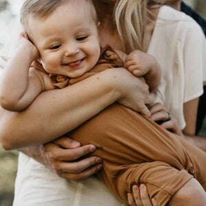 Smiling woman holding a baby in brown clothes outdoors.