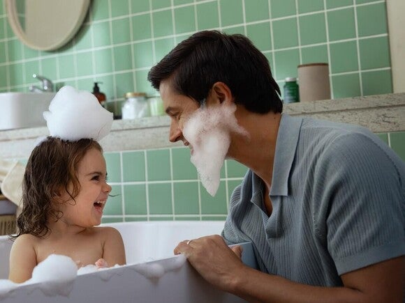 Child and adult with soap bubbles in a green-tiled bathroom.